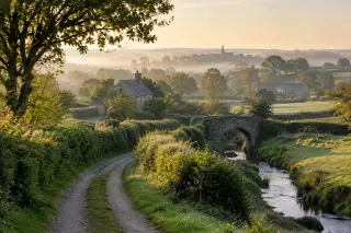Early morning rural landscape near Le Longeron with a dirt road, hedgerows, rolling fields, a stone farmhouse, a stream with a bridge, and a misty village in the distance under golden sunlight.